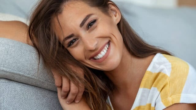A woman with long brown hair and a yellow striped shirt smiles brightly while leaning her head on a gray sofa.