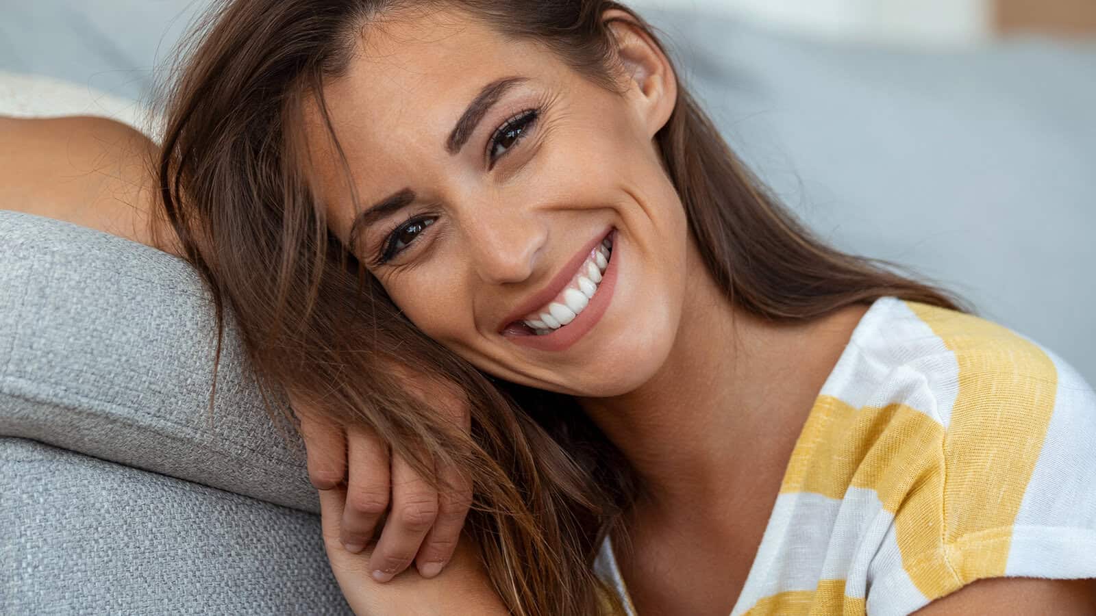 A woman with long brown hair and a yellow striped shirt smiles brightly while leaning her head on a gray sofa.