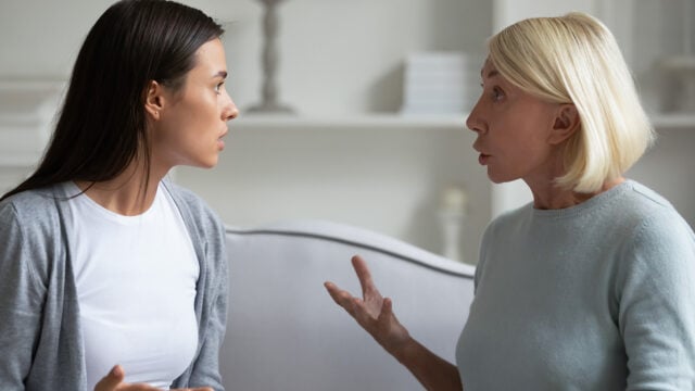 Two women sitting indoors on a couch are facing each other and appear to be having an intense or emotional conversation, with expressive hand gestures and serious facial expressions.