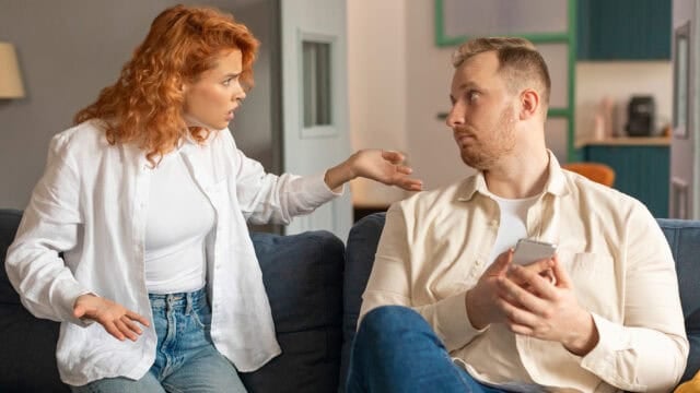 A woman gestures emotionally toward a man holding a smartphone, both sitting on a couch. The man looks away, appearing upset or defensive, while the woman seems frustrated. The setting is a modern living room.
