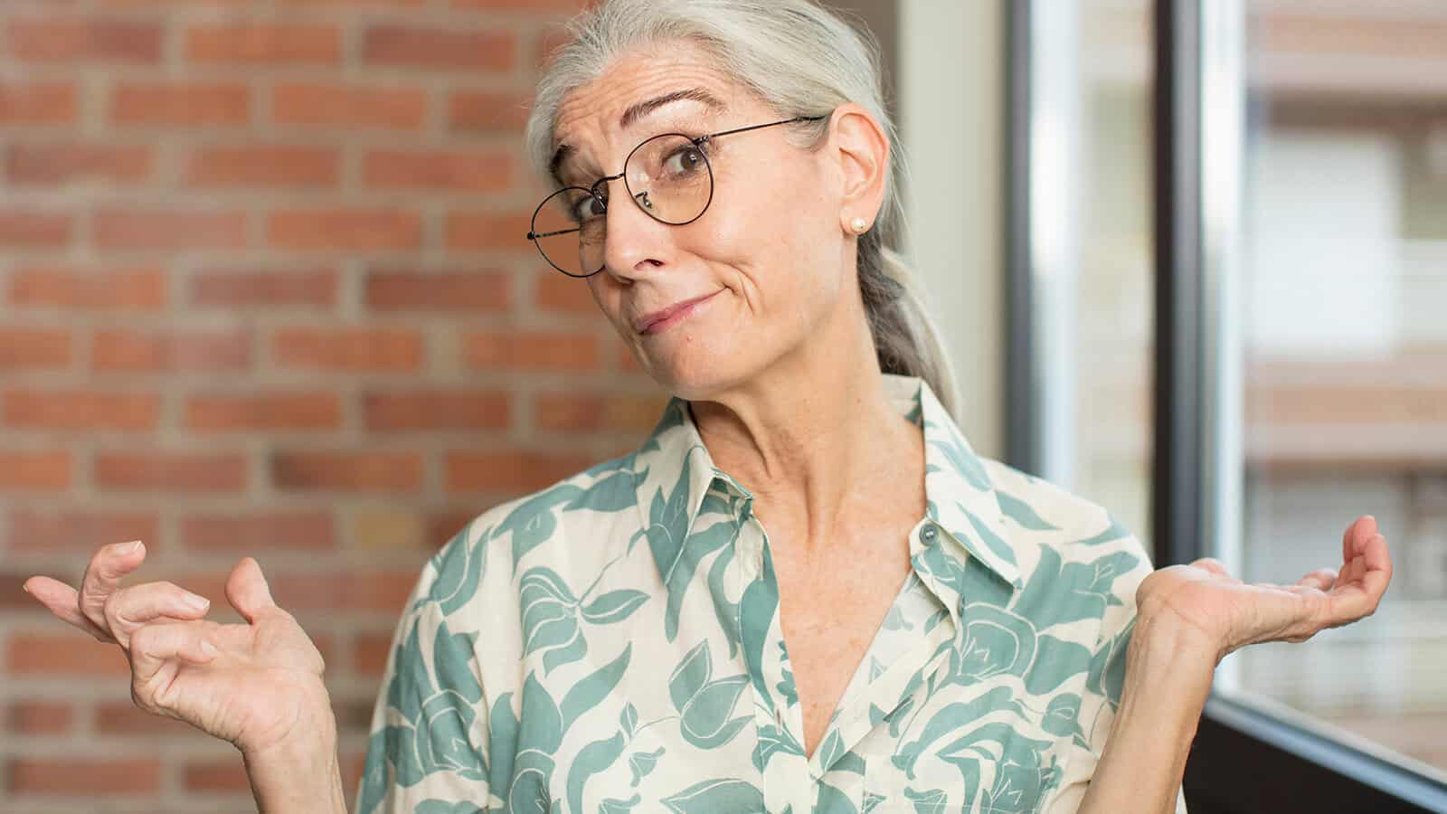 Older woman with gray hair, glasses, and a patterned shirt stands indoors, shrugging with both hands raised, and displaying a puzzled or uncertain expression. A brick wall and window are in the background.