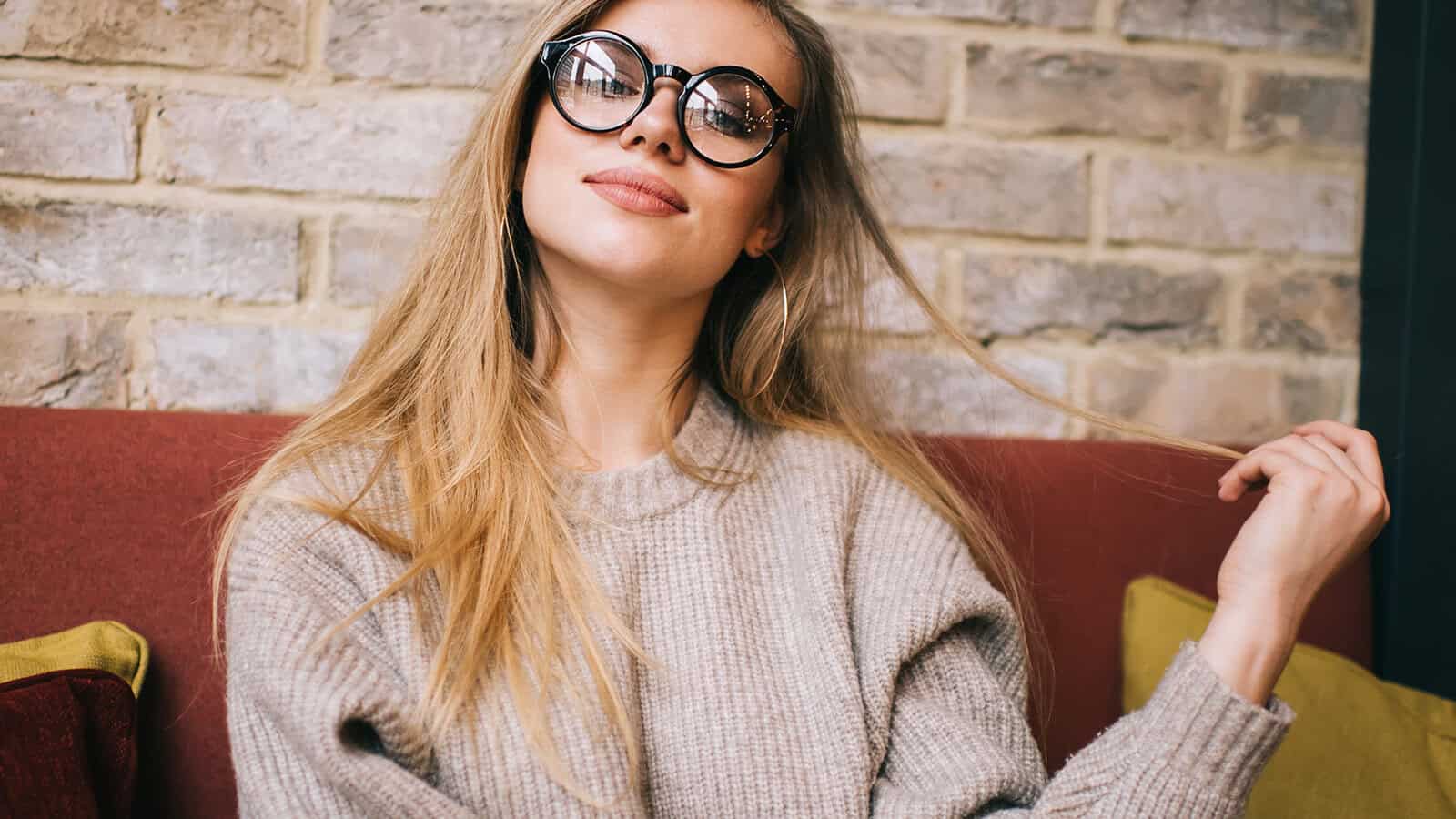 A woman with long blonde hair and round glasses sits on a red couch, wearing a beige sweater and smiling slightly while holding a strand of her hair. There is a brick wall in the background.