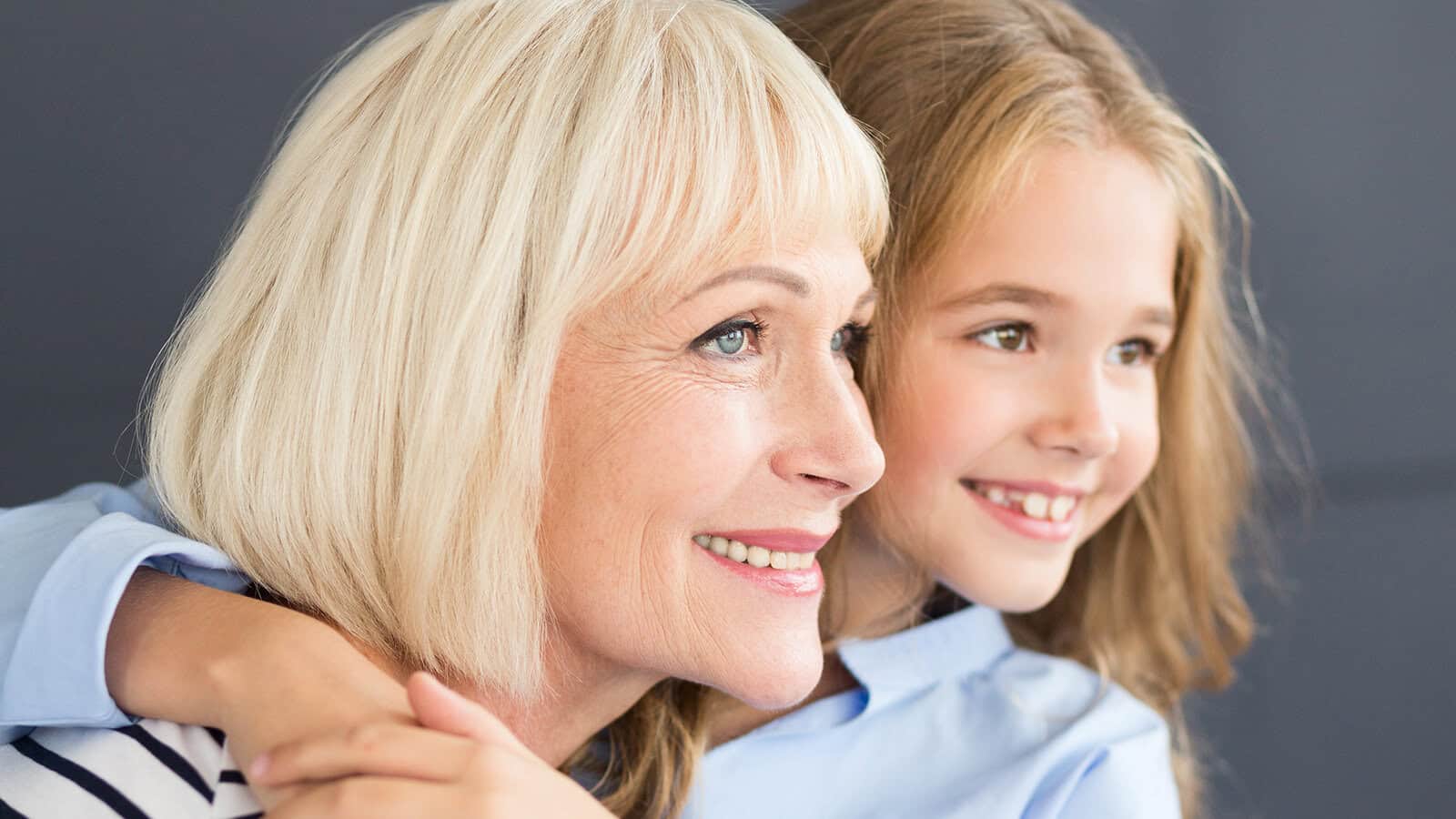 An older woman with short blonde hair smiles as a young girl with long light brown hair hugs her from behind. Both are looking slightly to the right, appearing happy and content.