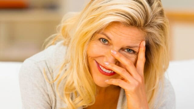 Smiling woman with long blonde hair looks at the camera, holding her hand partially over her face. She appears cheerful and relaxed, sitting indoors in a softly lit setting.
