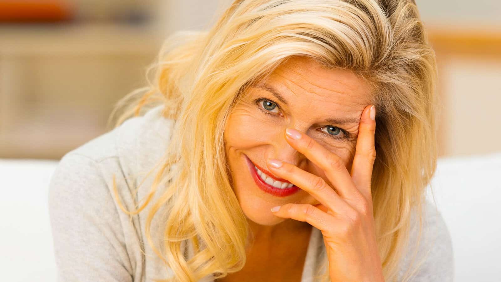 Smiling woman with long blonde hair looks at the camera, holding her hand partially over her face. She appears cheerful and relaxed, sitting indoors in a softly lit setting.