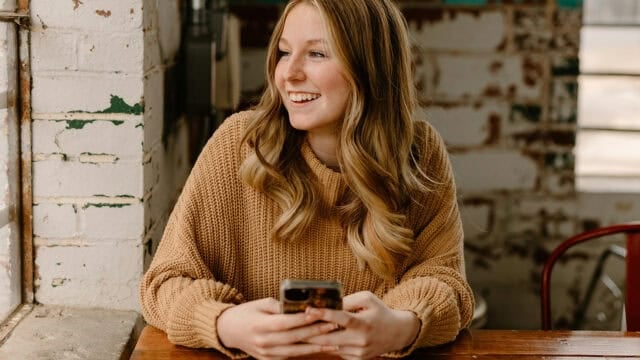 A woman with long blonde hair wearing a brown sweater sits at a wooden table, smiling and holding a smartphone. She looks out of a window in a rustic, sunlit cafe with exposed brick walls.