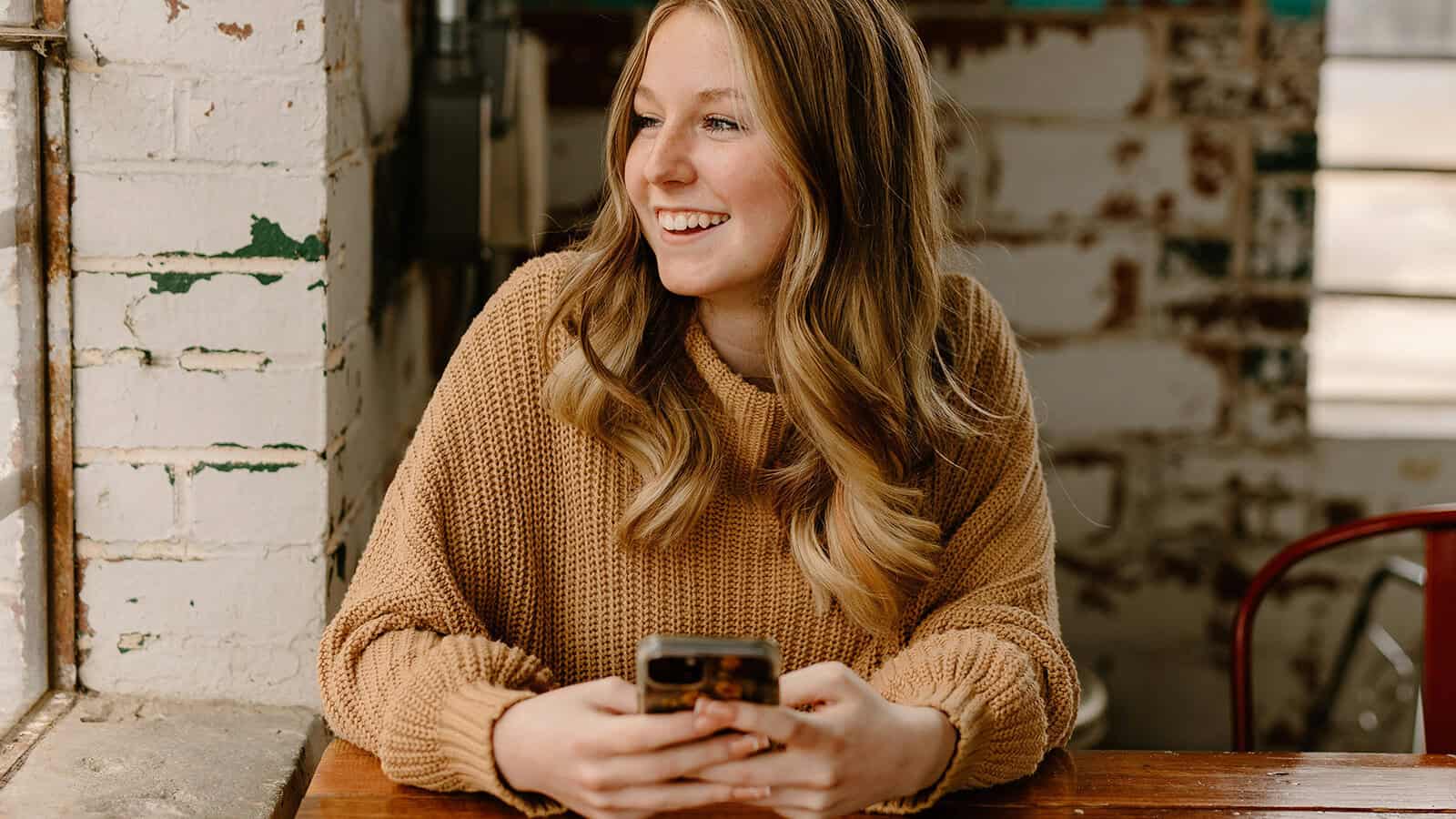 A woman with long blonde hair wearing a brown sweater sits at a wooden table, smiling and holding a smartphone. She looks out of a window in a rustic, sunlit cafe with exposed brick walls.