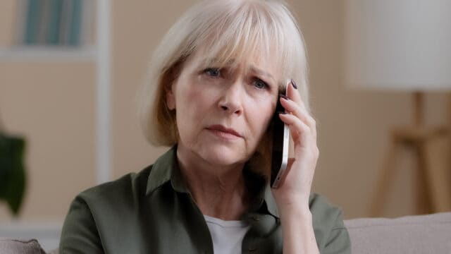 An older woman with short white hair looks worried while talking on a smartphone, sitting indoors.
