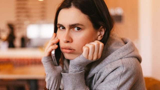 A person in a gray hoodie looks concerned while holding a phone to their ear, sitting indoors with a blurry background.