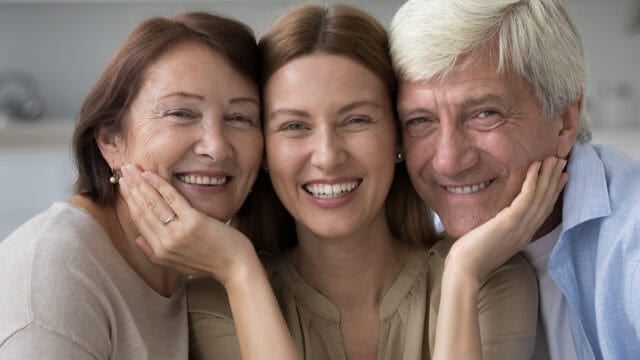 Three people smile warmly, their faces close together. The two older adults, one with gray hair and one with brown hair, embrace a younger woman in the middle, all looking happy and relaxed.