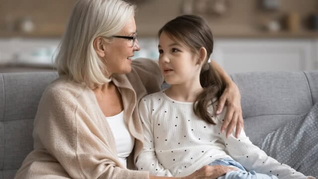 An older woman with gray hair and glasses sits on a couch, smiling and hugging a young girl with brown hair in a ponytail. They look at each other warmly in a cozy living room.