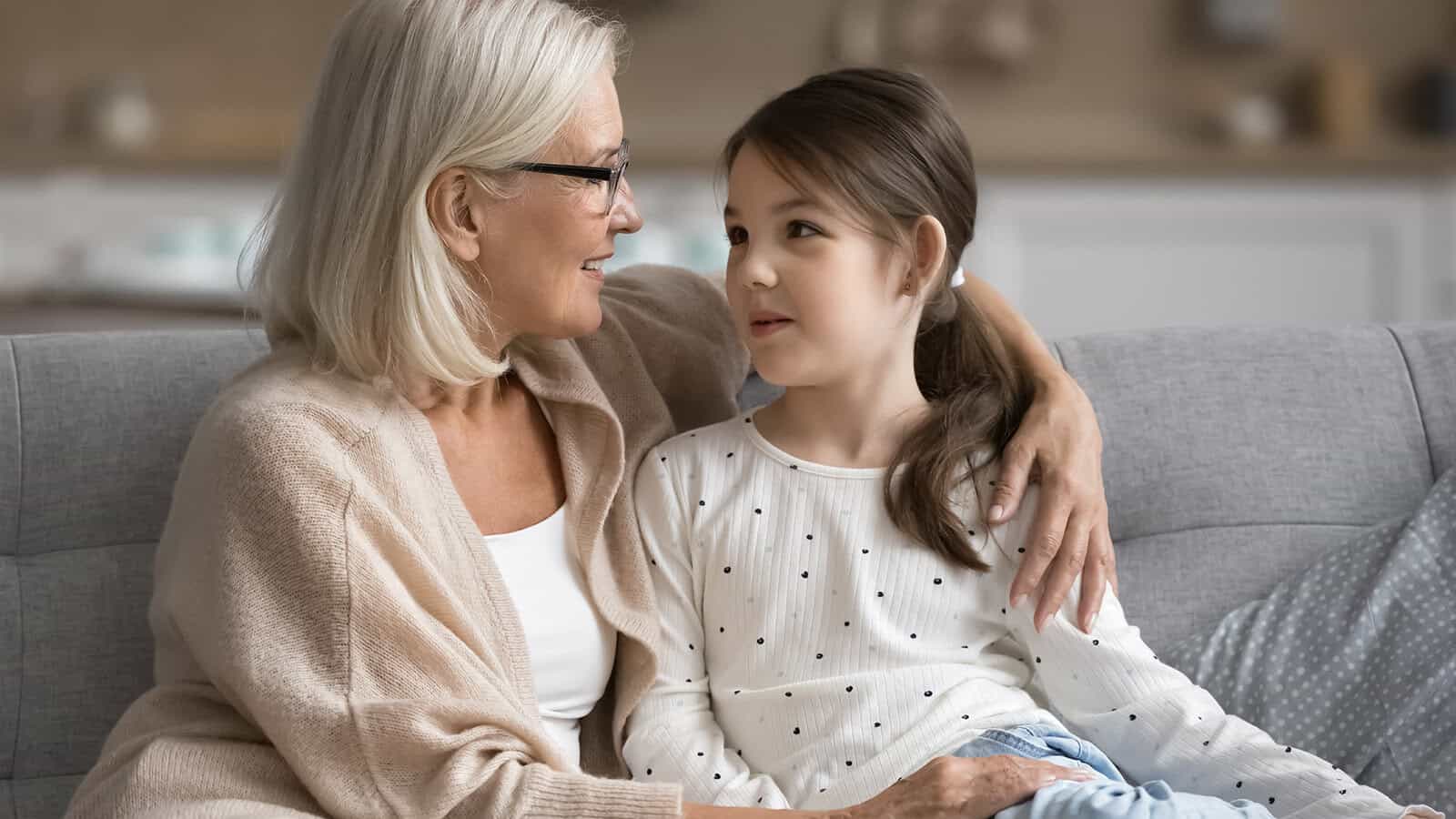An older woman with gray hair and glasses sits on a couch, smiling and hugging a young girl with brown hair in a ponytail. They look at each other warmly in a cozy living room.