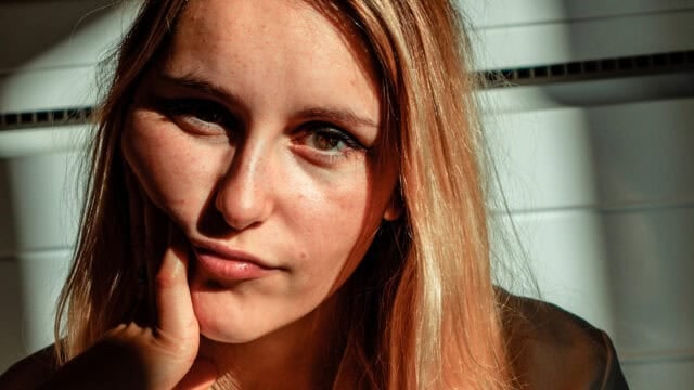 A young woman with long blonde hair rests her cheek on her hand, gazing at the camera in sunlight, with shadows falling across her face and a neutral background.