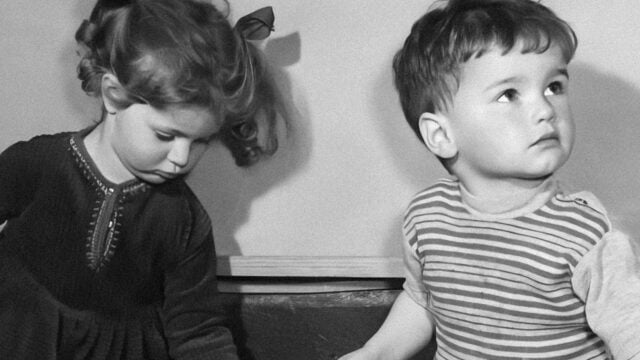 Black and white photo of a young girl with curly hair and bows looking down, next to a young boy in a striped shirt looking up and to the right. Both children appear thoughtful and are sitting close together indoors.