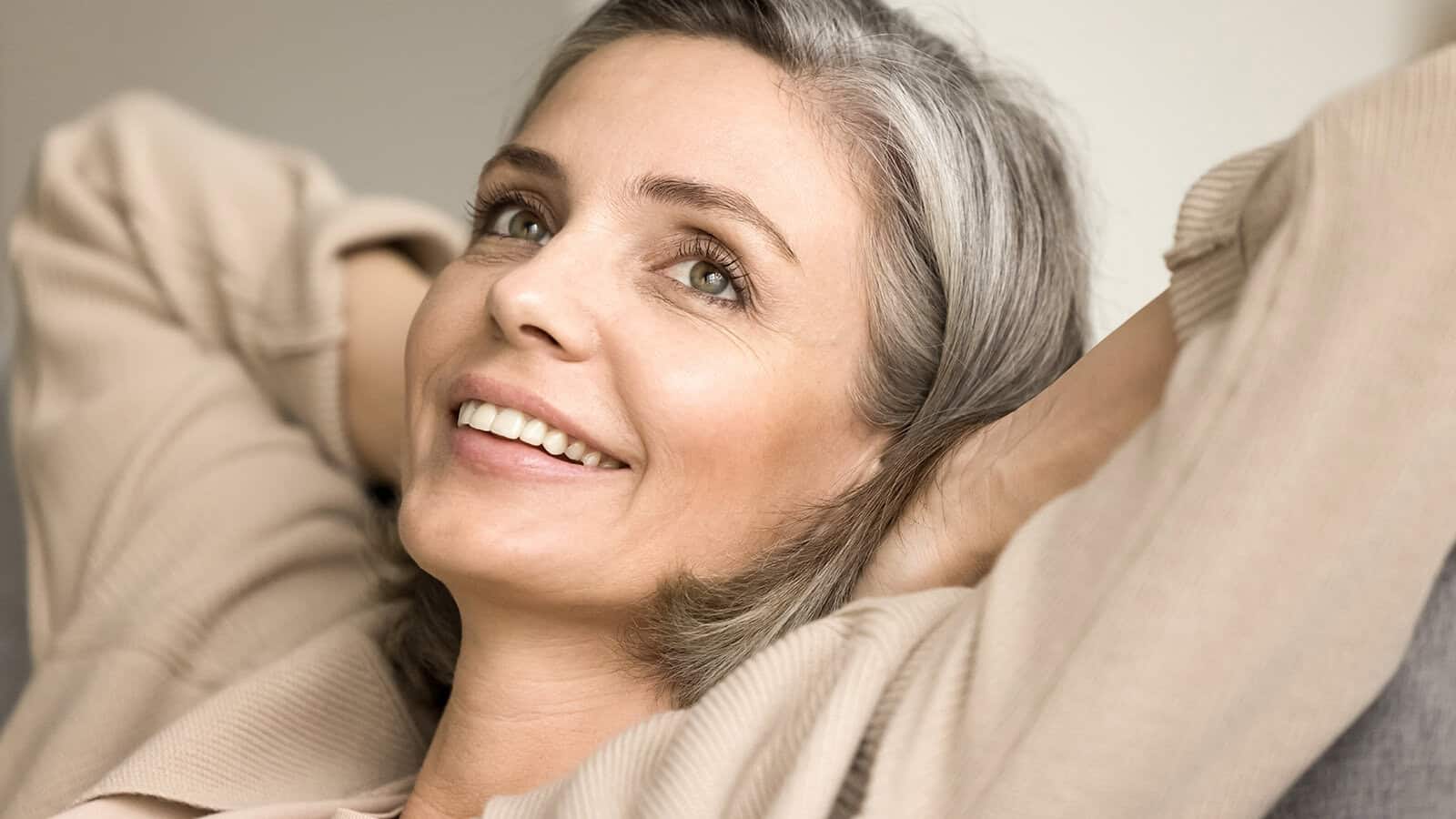 A woman with gray hair smiles gently while relaxing with her arms behind her head, wearing a beige top and looking content.