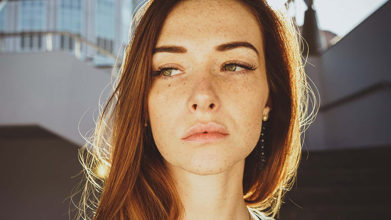 A young woman with long red hair and freckles looks to the side in sunlight, with blurred urban buildings and stairs in the background.