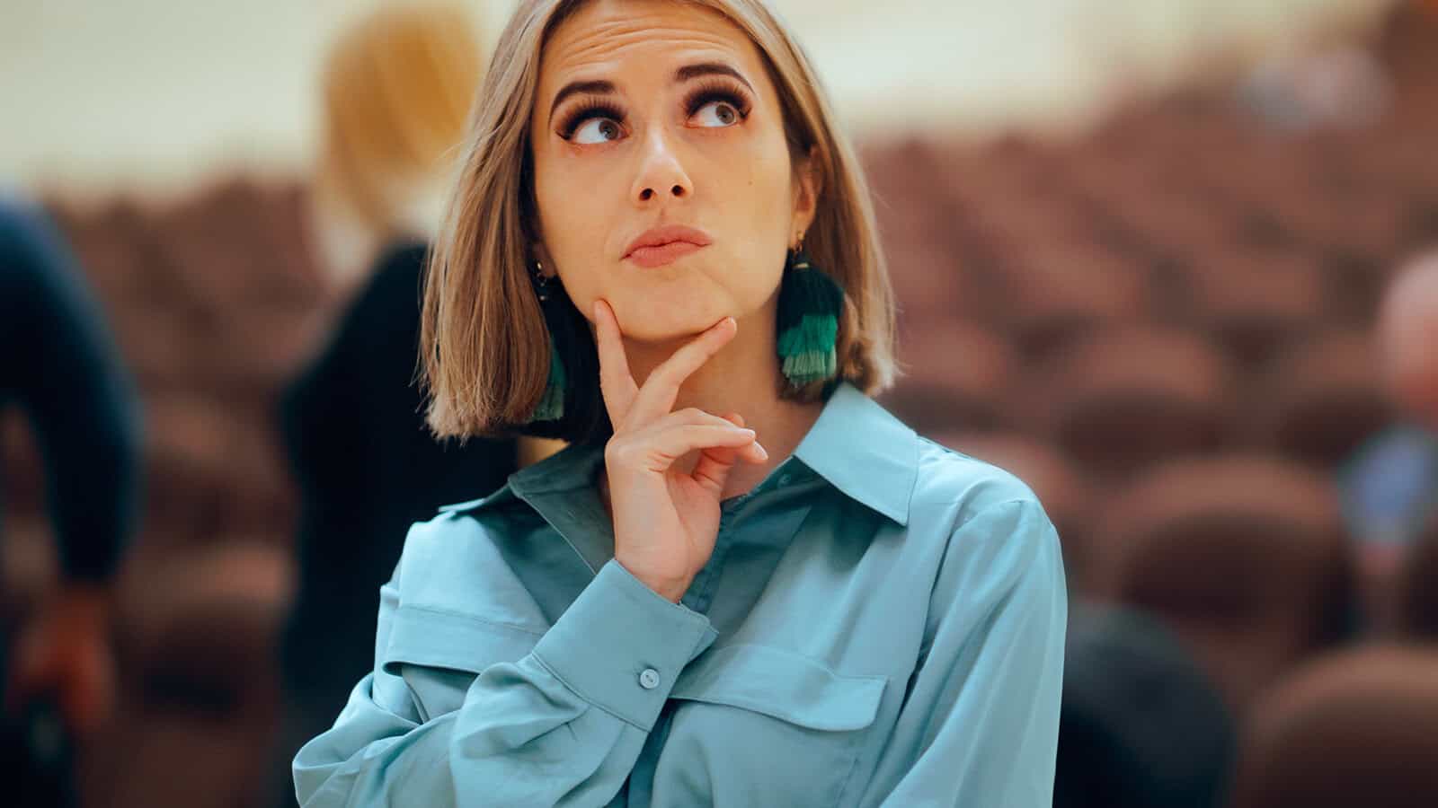 A woman with short hair and large earrings looks up thoughtfully with her hand on her chin, standing in a blurred indoor setting with empty rows of chairs.