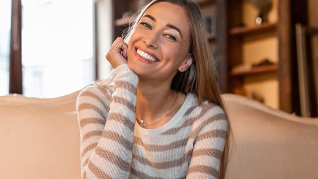 A woman with long hair in a striped sweater sits on a couch, smiling brightly at the camera in a warmly lit room with shelves in the background.