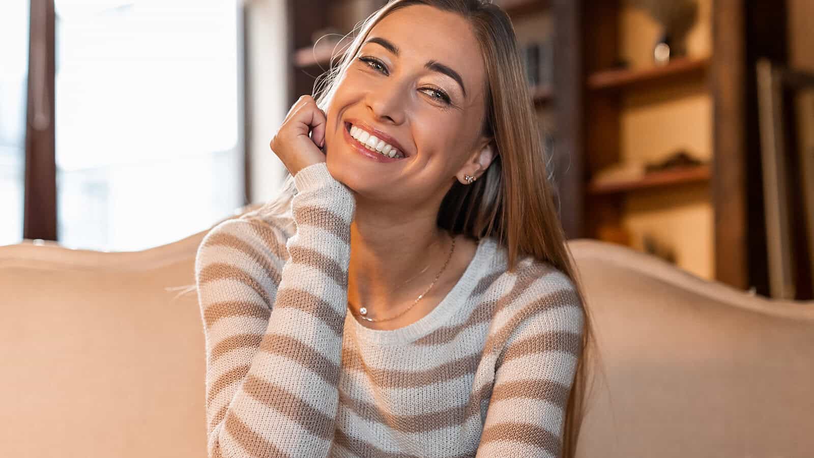A woman with long hair in a striped sweater sits on a couch, smiling brightly at the camera in a warmly lit room with shelves in the background.