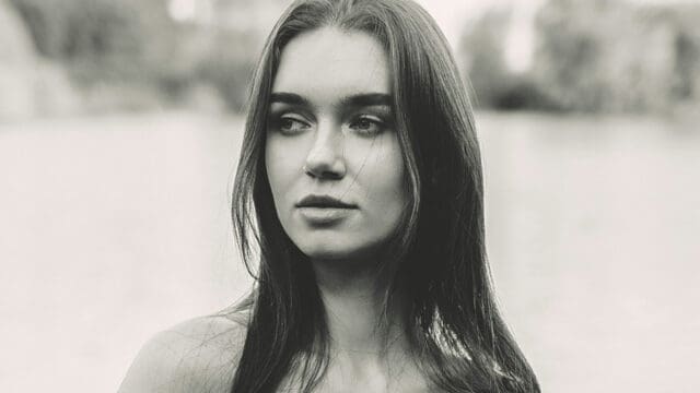 A young woman with long, straight hair looks to the side with a neutral expression. The image is in black and white with a blurred outdoor background.