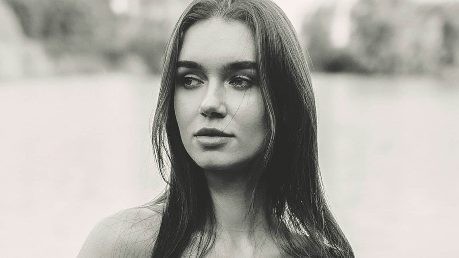 A young woman with long, straight hair looks to the side with a neutral expression. The image is in black and white with a blurred outdoor background.