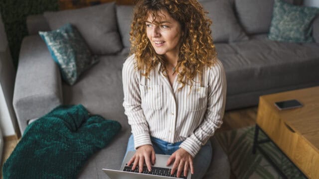 A woman with curly hair sits on a gray sofa using a laptop, looking to the side and smiling. A green blanket and patterned pillows are on the couch, with a phone on a wooden table nearby.