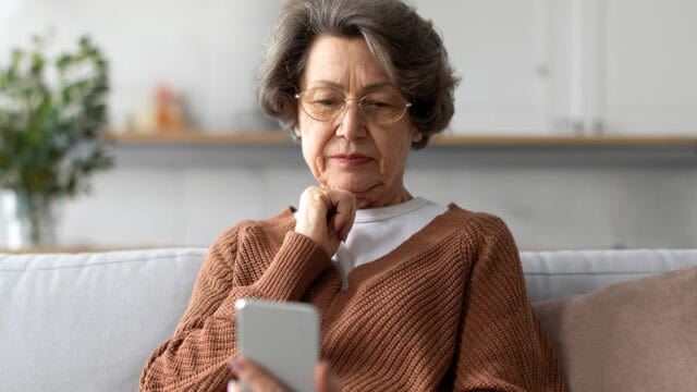 An older woman with gray hair and glasses sits on a couch, wearing a brown sweater, thoughtfully looking at a smartphone in her hand.
