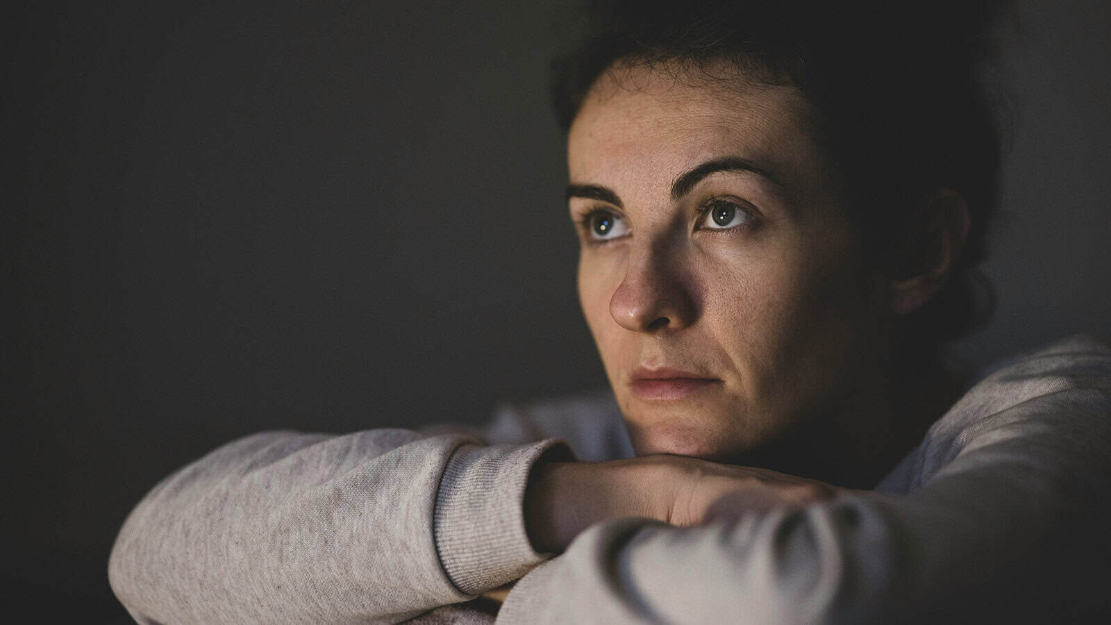 A person with dark hair looks thoughtful and serious, resting their chin on their arms, gazing slightly upward in dim lighting.