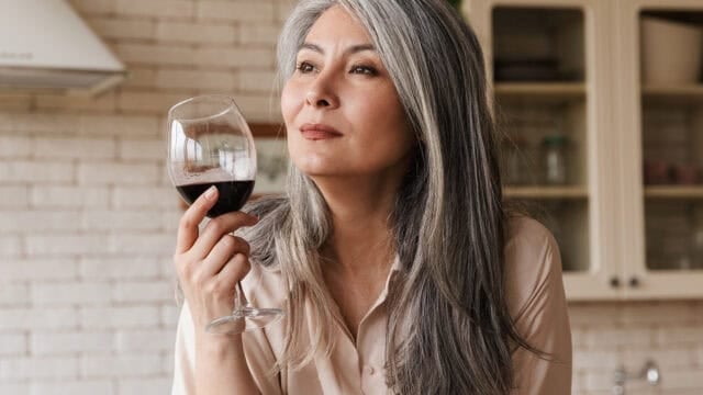A woman with long gray hair holds a glass of red wine, looking thoughtful. She is indoors, and kitchen cabinets are visible in the background.