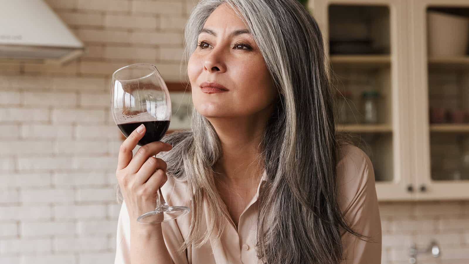 A woman with long gray hair holds a glass of red wine, looking thoughtful. She is indoors, and kitchen cabinets are visible in the background.