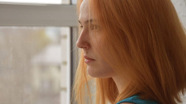 A woman with long red hair looks thoughtfully out of a window, contemplating the nuances of social perception and the elusive nature of the liking gap.