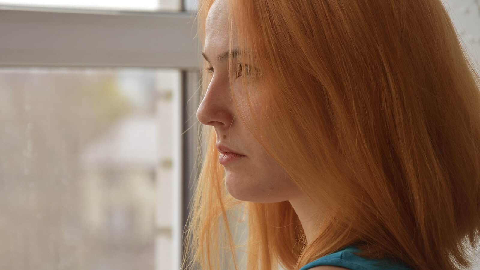 A woman with long red hair looks thoughtfully out of a window, contemplating the nuances of social perception and the elusive nature of the liking gap.