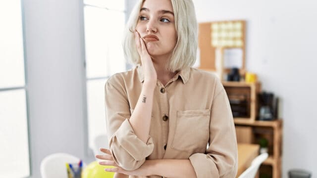 A person with short blonde hair and a beige shirt stands indoors, resting their chin on one hand and looking thoughtful or bored.