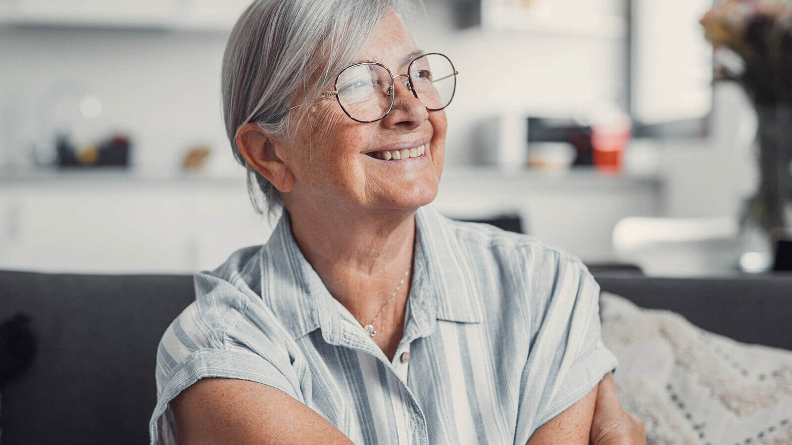 Smiling older woman with gray hair and glasses sits on a couch in a bright room, wearing a light-colored, short-sleeve, striped shirt.