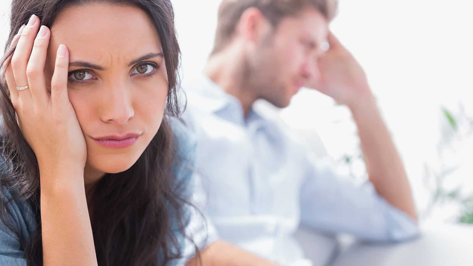A woman looks upset with her hand on her face, while a man sits in the background, appearing stressed and looking away.