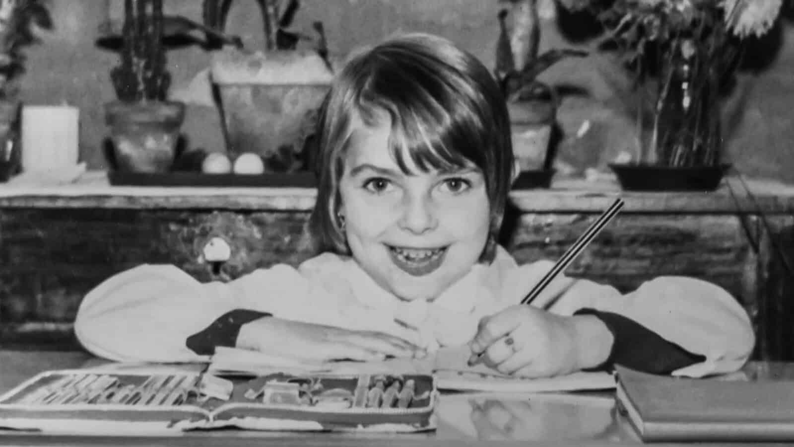 A young girl smiles at the camera while drawing with colored pencils at a desk. Plants and books are visible in the background.