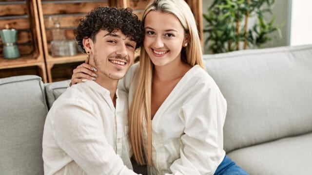 A young couple sits closely together on a light gray couch, smiling at the camera. The man has curly dark hair and the woman has long blonde hair. Both are wearing white shirts. Wooden shelves and plants are in the background.