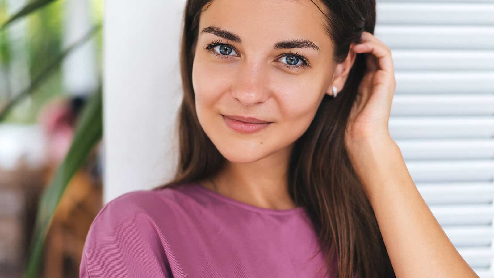 A young woman with long brown hair and blue eyes, wearing a mauve shirt, gently touches her hair and smiles softly at the camera. She stands indoors near a white wall and partially open window shutters.