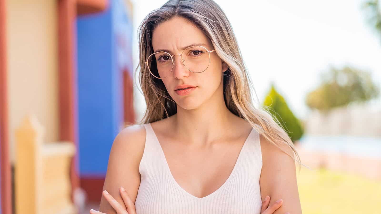 A woman with long hair and glasses stands outdoors with arms crossed, wearing a white tank top and looking at the camera with a serious, slightly confused expression.