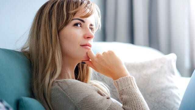 Woman with long blonde hair sits on a sofa, resting her chin on her hand and gazing thoughtfully into the distance. Neutral-toned background with soft natural light.