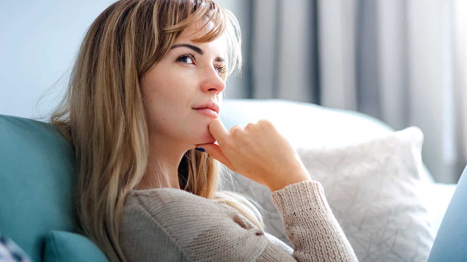 Woman with long blonde hair sits on a sofa, resting her chin on her hand and gazing thoughtfully into the distance. Neutral-toned background with soft natural light.