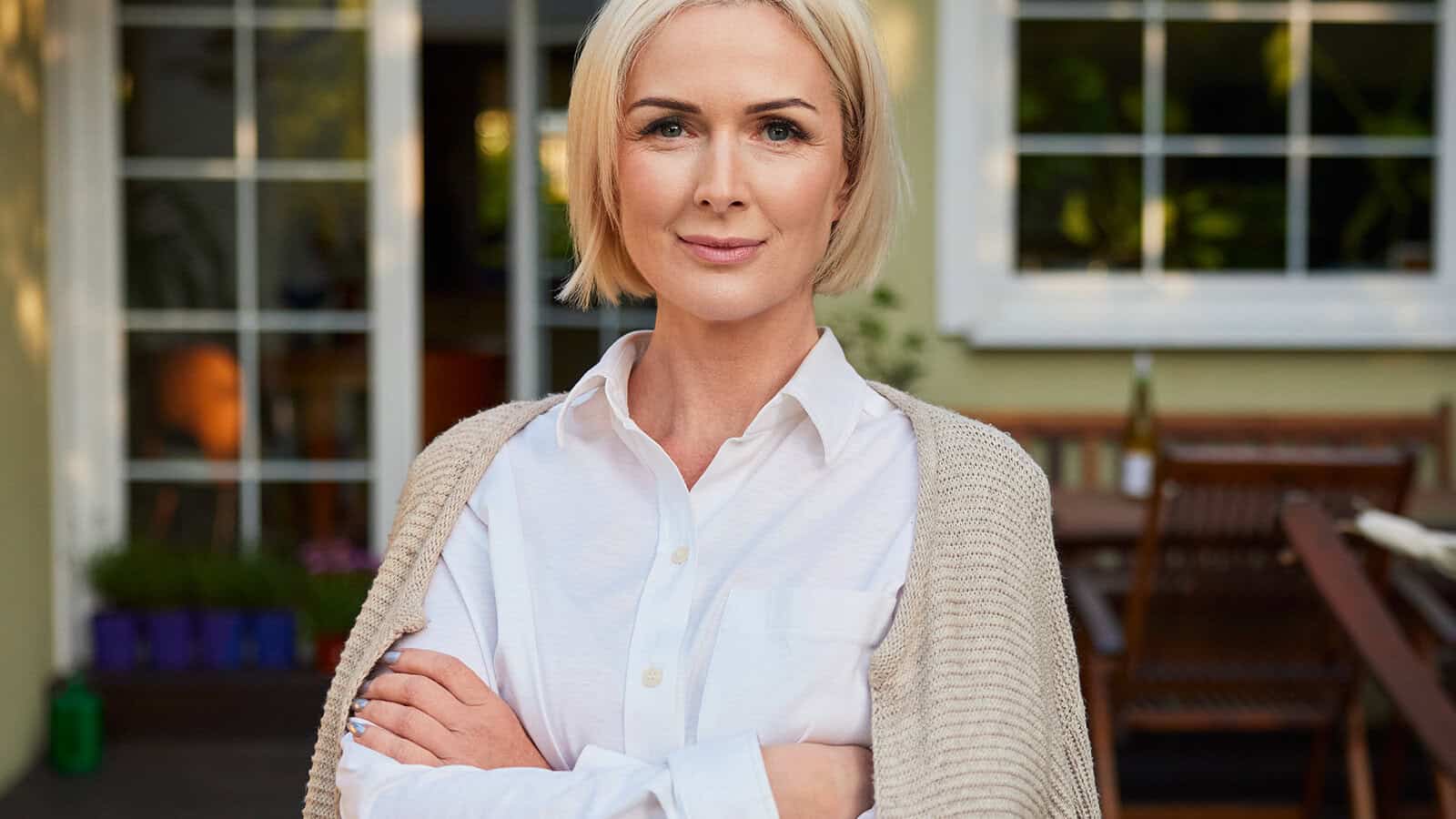 A woman with short blonde hair stands outside in front of a house, wearing a white shirt and beige sweater, arms crossed, looking confidently at the camera.