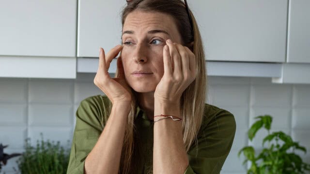 A woman with long hair stands in a kitchen, using both hands to insert a contact lens into her right eye. She is wearing a green shirt and has a bracelet on her left wrist.