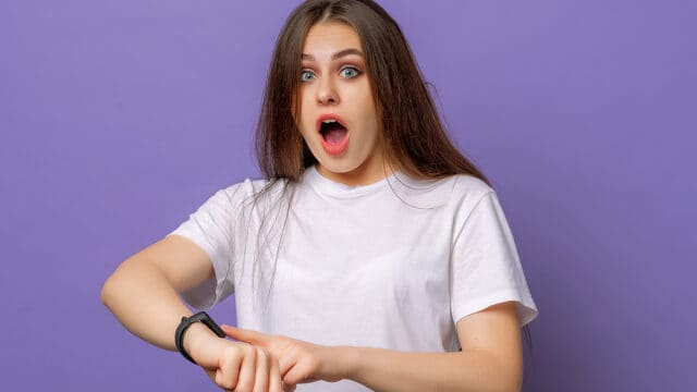 A young woman with long brown hair wearing a white t-shirt looks surprised while checking the time on her smartwatch, standing in front of a purple background.