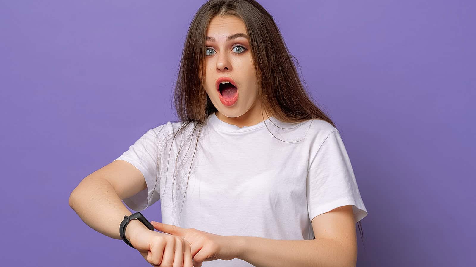 A young woman with long brown hair wearing a white t-shirt looks surprised while checking the time on her smartwatch, standing in front of a purple background.
