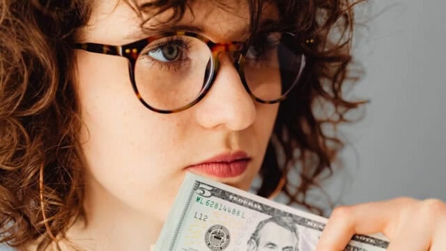 A person with curly hair and glasses holds a five-dollar bill close to their face, looking thoughtful.