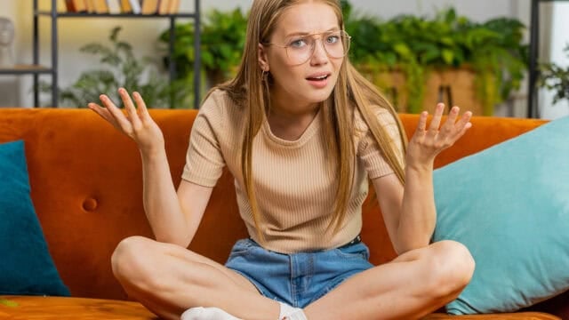 A young Gen Z woman wearing glasses sits cross-legged on an orange sofa, shrugging with her hands up and looking confused. Green plants and pillows are in the background.