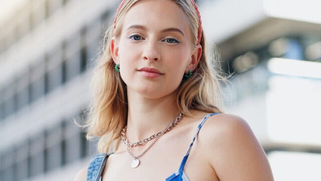 A young woman with blonde hair wears a red headband, layered necklaces, and a blue checkered top, standing outdoors with a blurred modern building in the background.