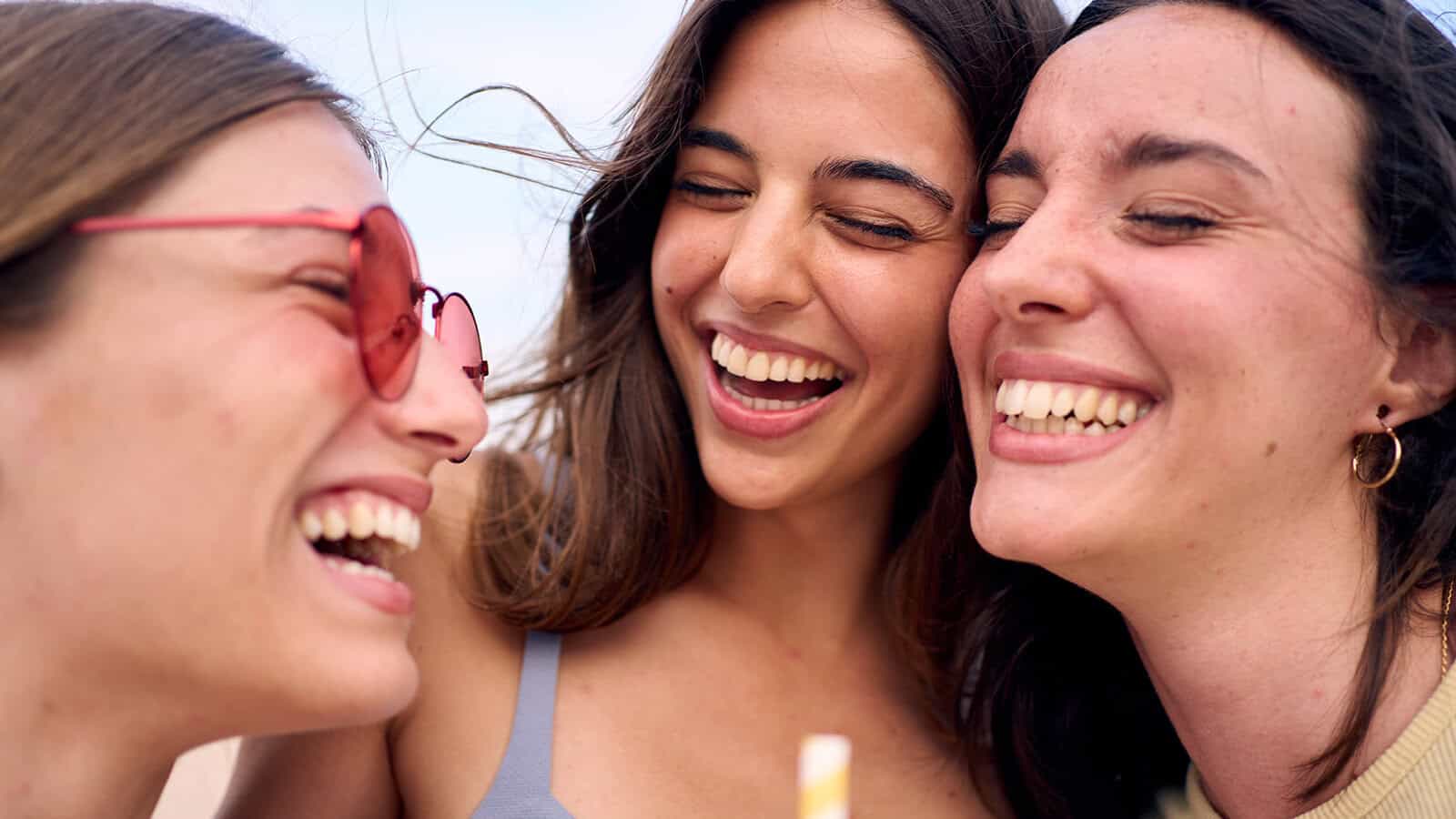Three young women smiling and laughing together, close up, outdoors. One wears pink sunglasses, and all appear joyful and relaxed, enjoying each others company.