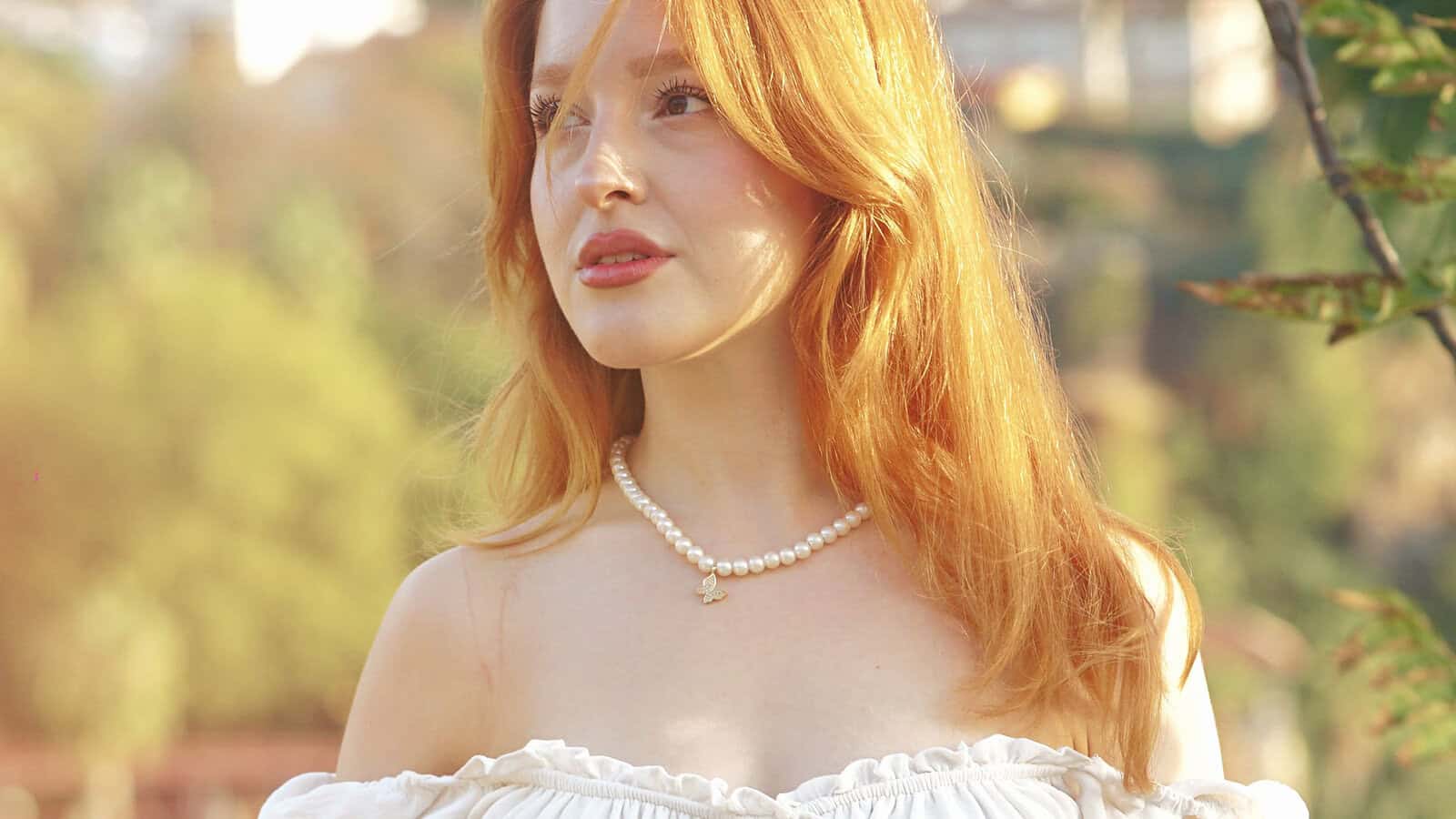 A woman with long red hair wears an off-shoulder white top and a pearl necklace, standing outdoors in soft sunlight with greenery in the blurred background.